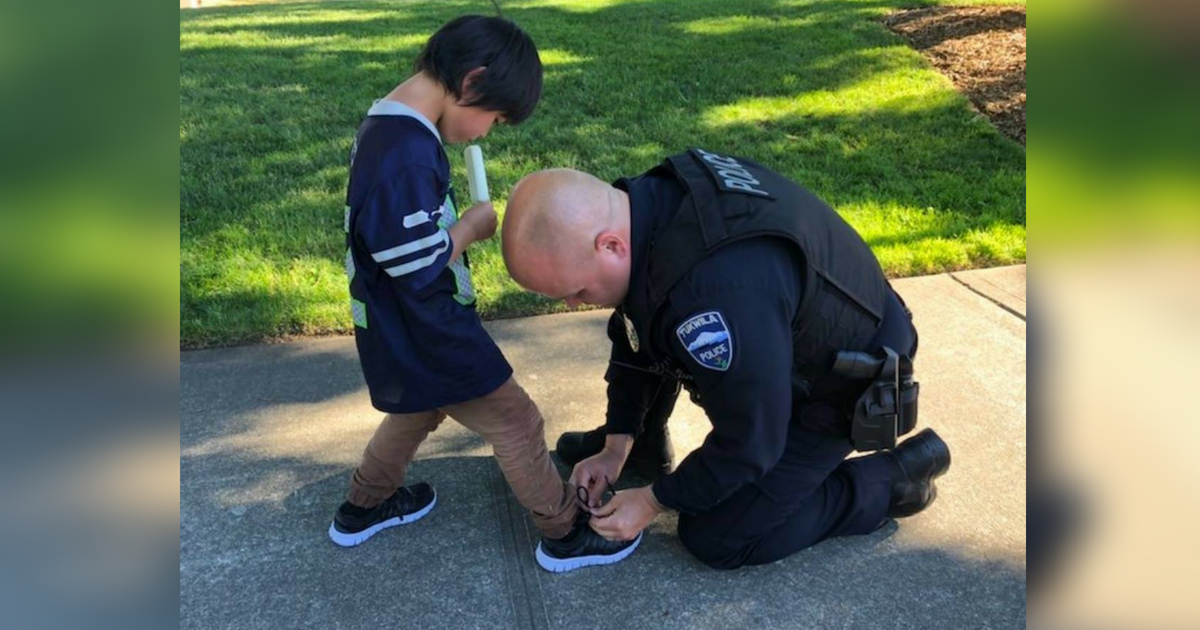 A cop kneels down to help put a new pair of shoes on a little boy after giving him a Popsicle.