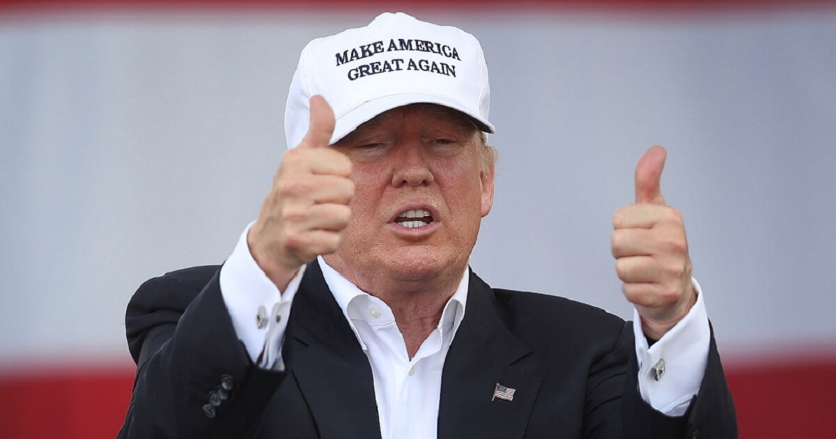 Donald Trump flashes two thumbs up while wearing a "Make America Great Again" baseball cap.