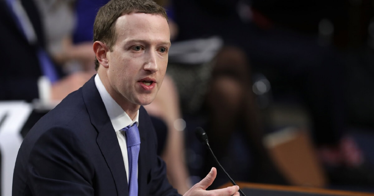 Mark Zuckerberg gestures while seated at a table before a microphone.