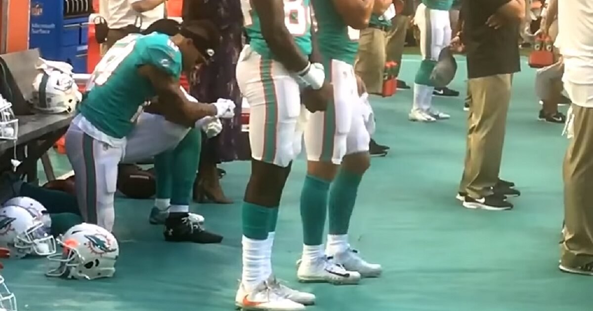 Football player kneels as teammates stand.