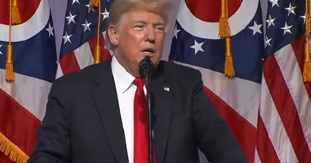 Trump at microphone with flags behind him.
