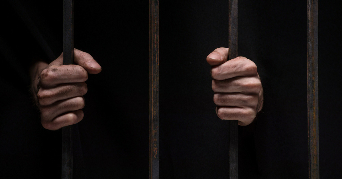 closeup on hands of man sitting in jail. Man behind jail bars on black background