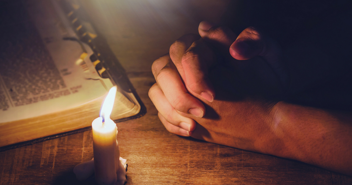 praying hands and blurred open bible on wooden table with candle light and light from above