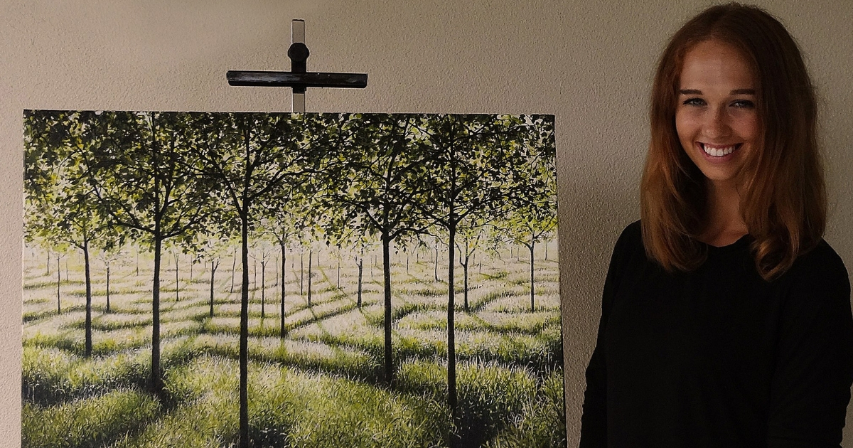 Girl stands next to her painting of a grove of trees.