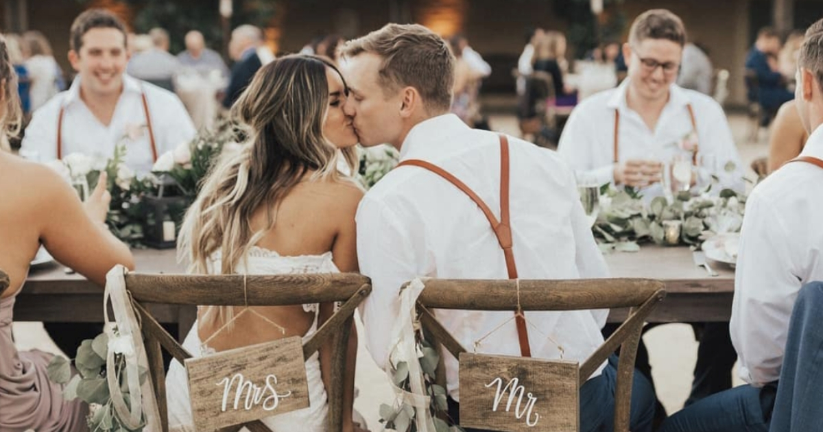 Newlyweds share a kiss while sitting in chairs labeled "Mrs." and "Mr."