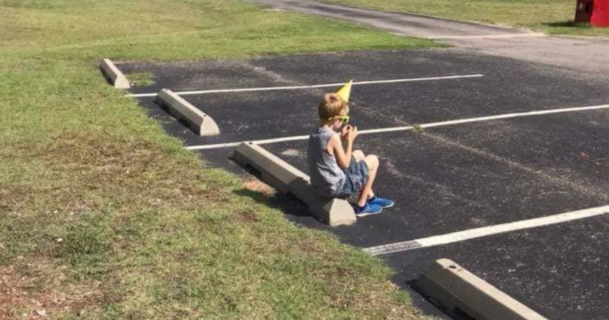 A little boy in a birthday hat sits alone in a parking lot.