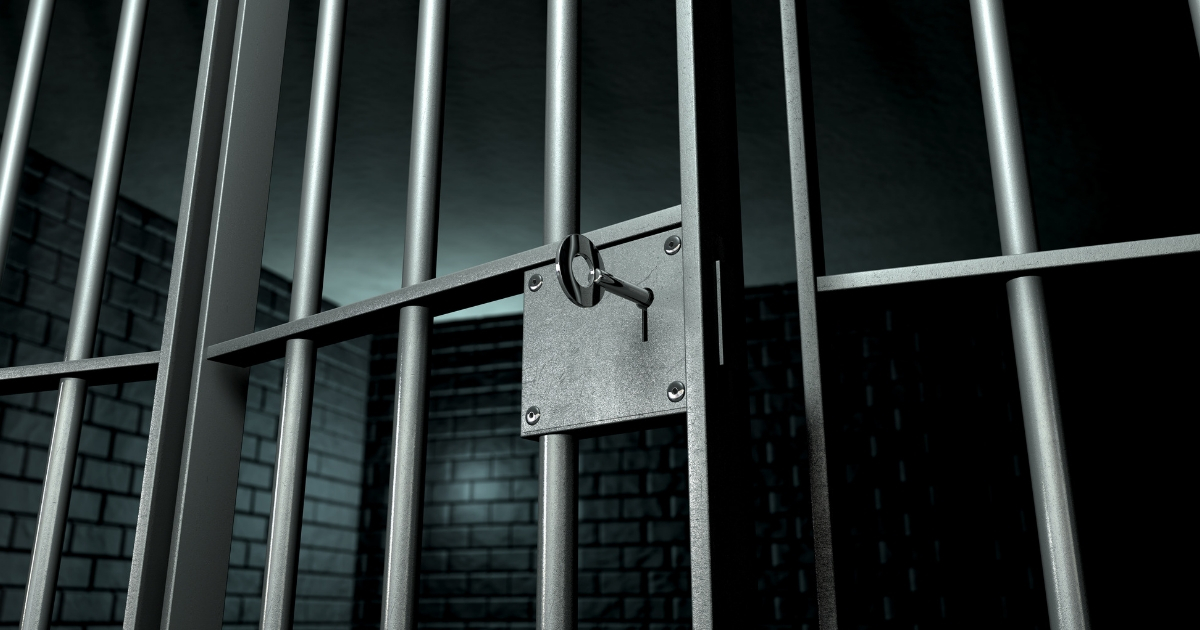 A closeup of the lock of a brick jail cell with iron bars and a key in the locking mechanism with the door open.