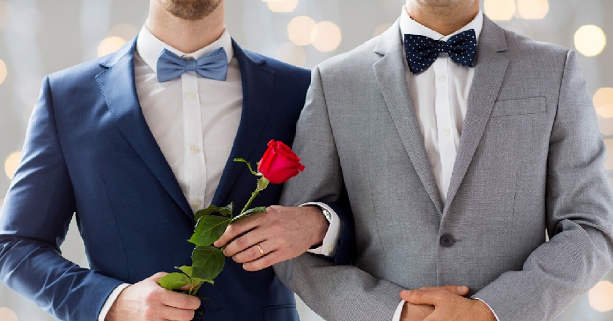 Two men in tuxedoes with their arms linked, one holding a rose.