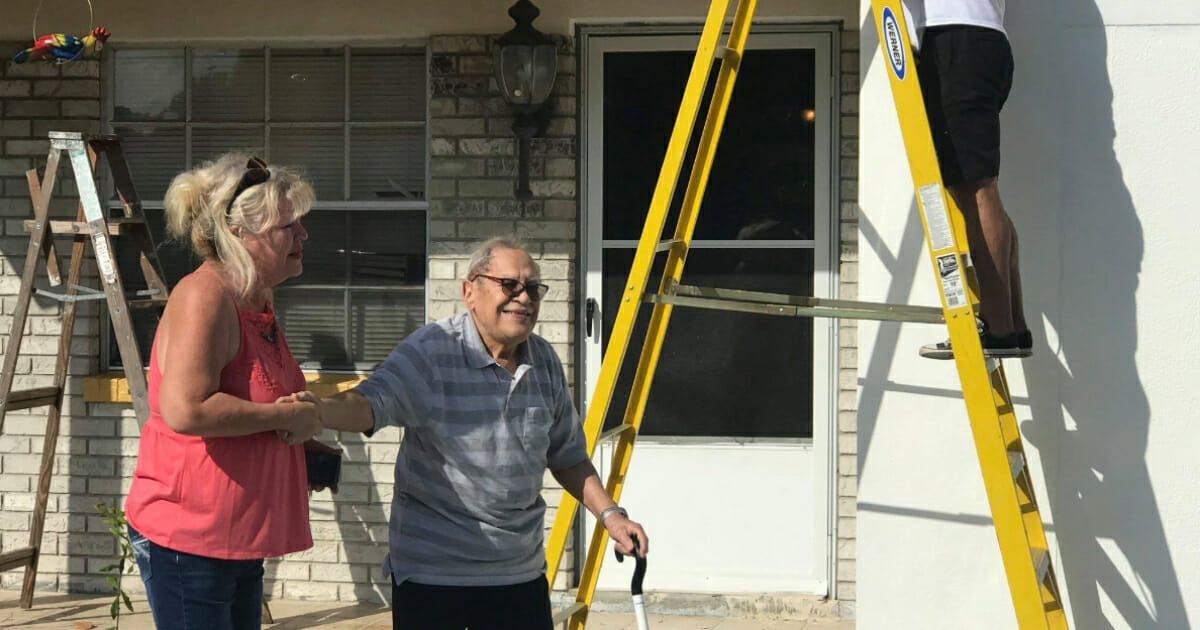 Maria Velez walks alongside her veteran husband as a man uses a ladder to paint the house