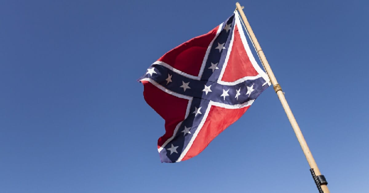 A Confederate flag flies from a pole against a blue sky.