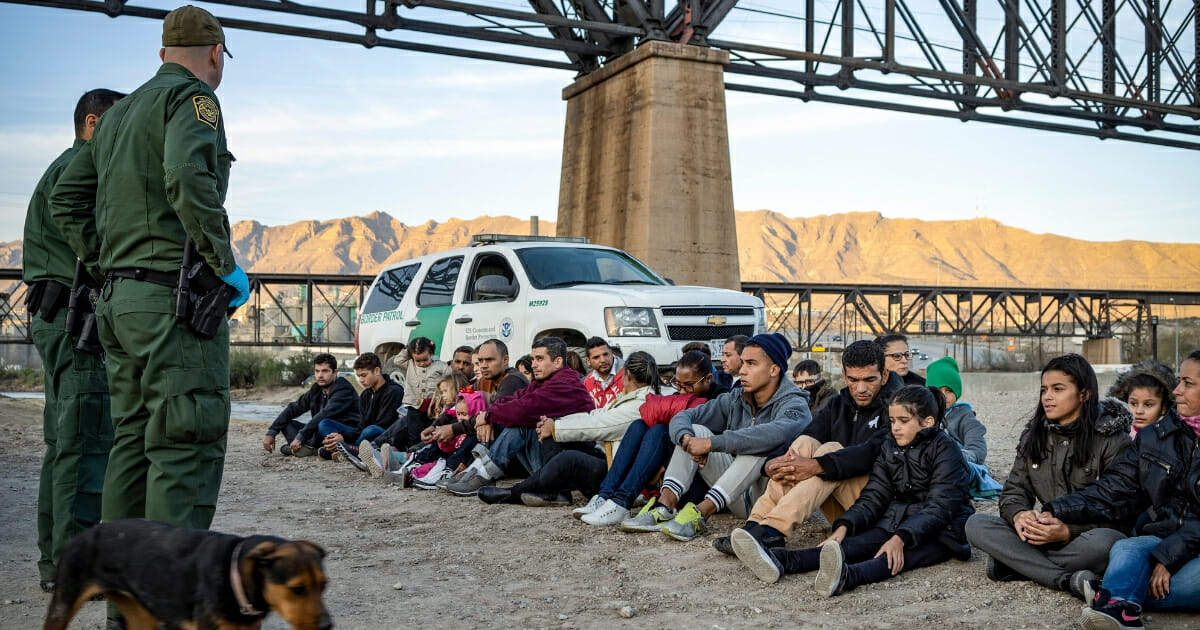 Border Patrol agents stand next to a large group of seated immigrants.