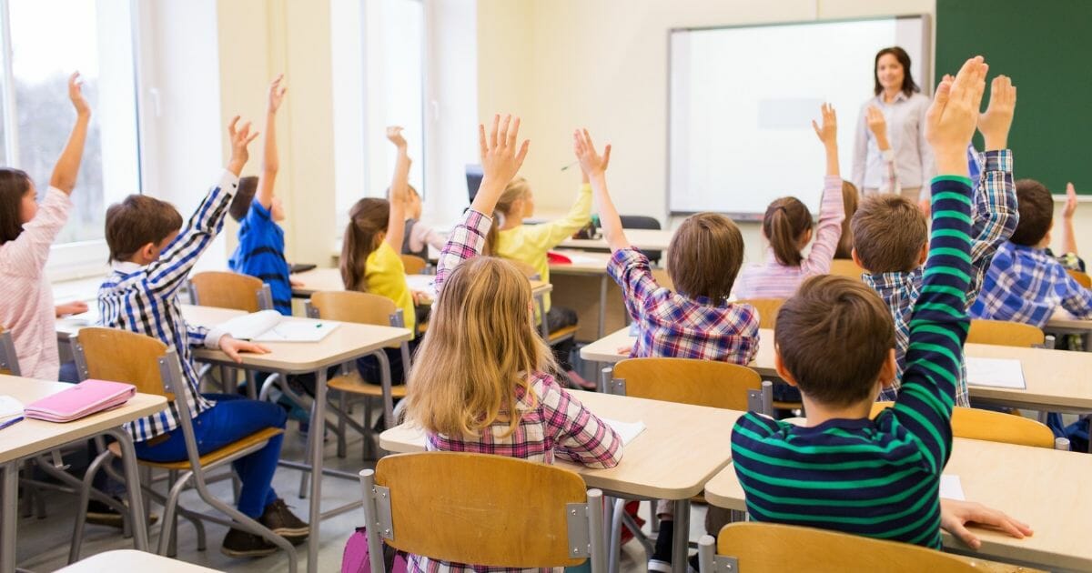 Group of school kids with teacher sitting in classroom and raising hands.