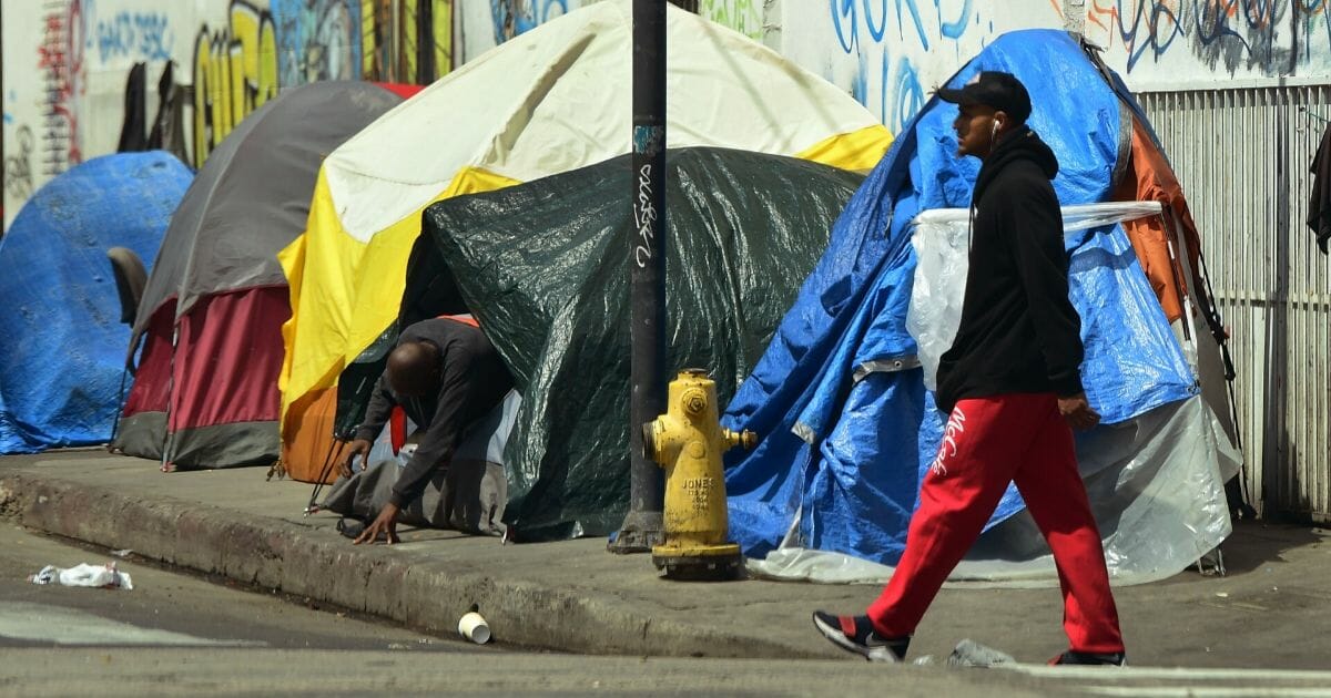 A pedestrian passes by as a man makes his way out of a tent among a row of tents on a sidewalk in downtown Los Angeles in May.