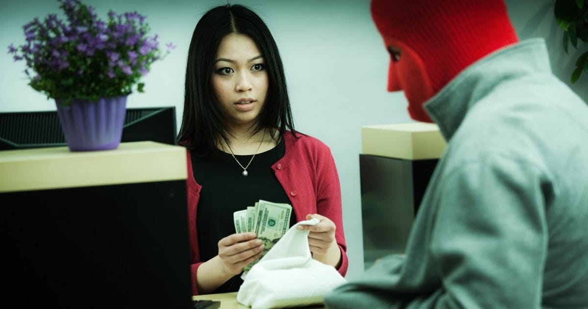 A robber wearing a ski mask and gray jacket watches as cash is being stuffed into a bag by a fearful bank teller across a retail bank counter.