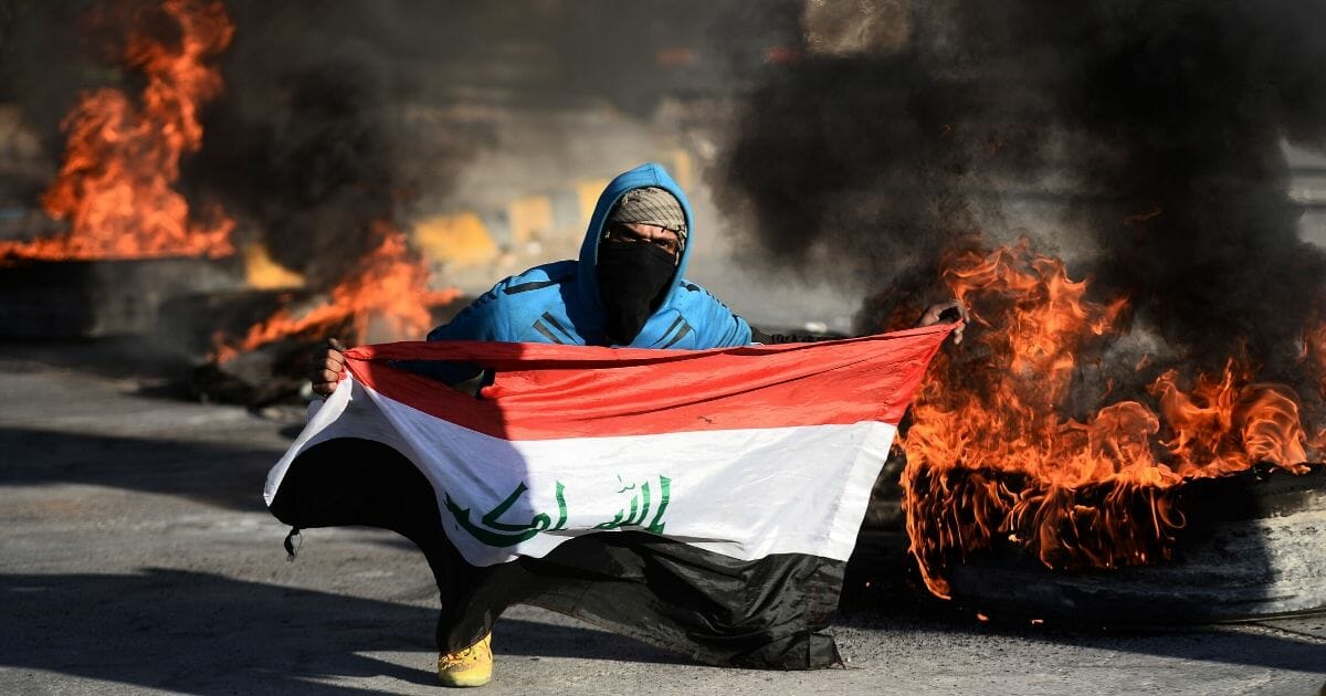 An Iraqi demonstrator carries a national flag while posing next to burning tires as angry protesters block roads in the central shrine city of Najaf on Jan. 5, 2020.