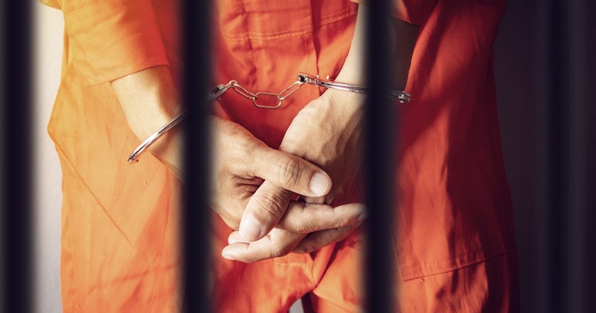 Cuffed hands seen in a rear view through jail bars.