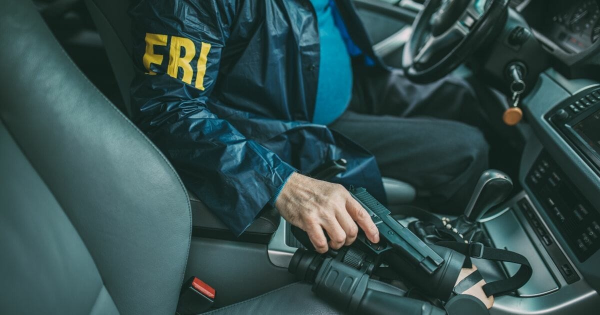 A man wearing an FBI jacket and holding a handgun sits at the wheel of a vehicle.