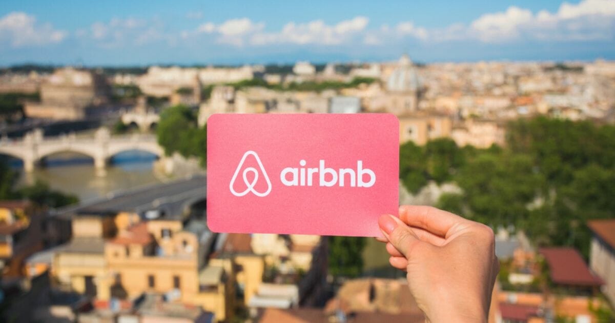 Stock image of a person holding a card displaying the Airbnb logo with Rome in the background.
