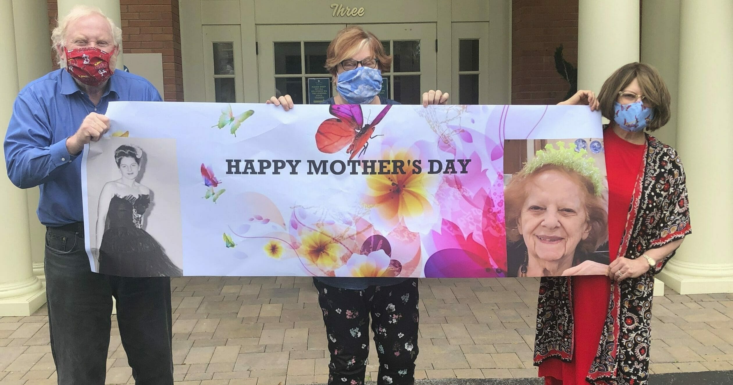 This May 3, 2020, photo released by Shelly Solomon shows, from left, Steve Turner and his sisters, Carla Paull and Lisa Fishman, holding up a Mother’s Day banner emblazoned with images of their mom, Beverly Turner, in front of her assisted living facility in Ladue, Missouri.