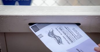 Stock image of a person depositing a mail-in ballot into a drop-off box.