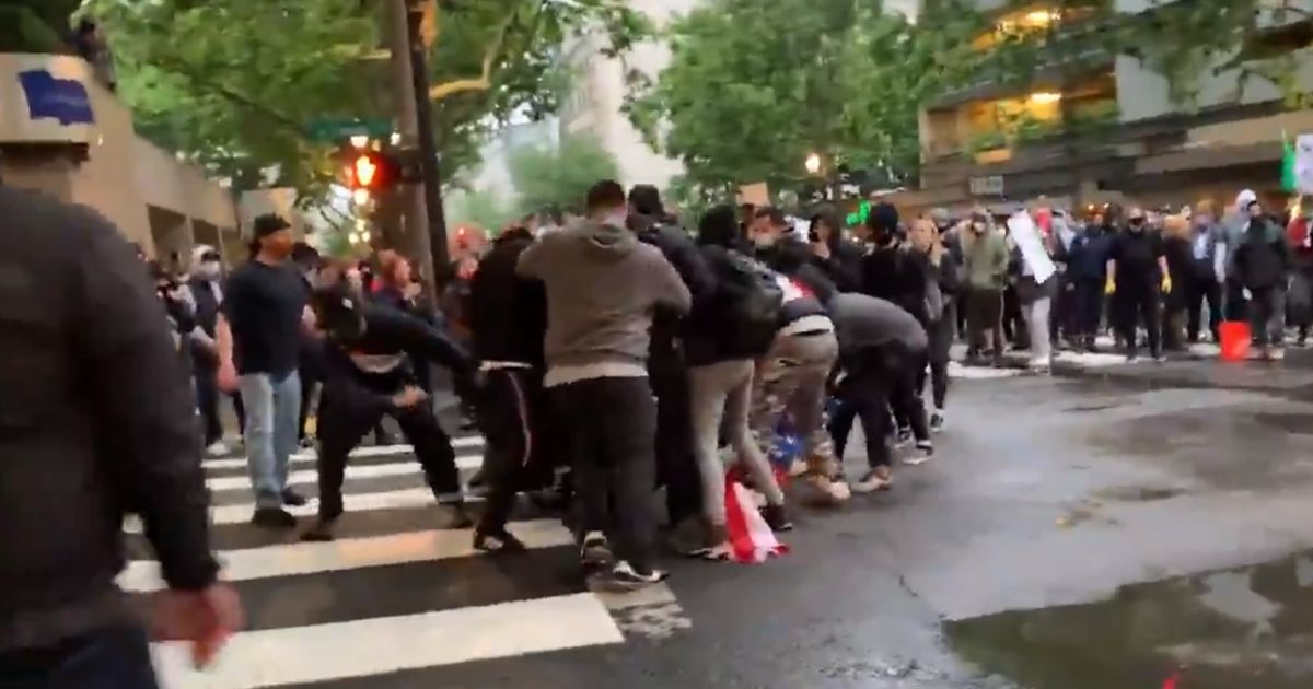 Rioters in Portland, Oregon, swarm a man clinging to an American flag.