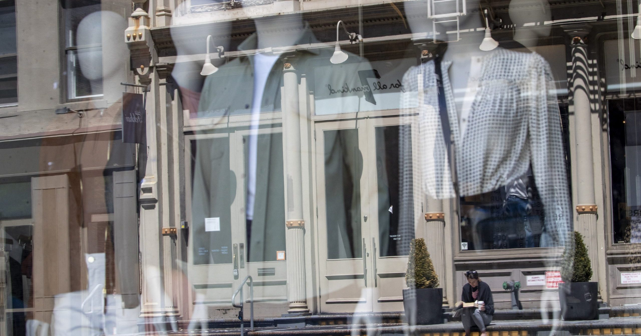 In this May 7, 2020 file photo a woman sitting on a stoop reading a book in the sun is seen reflected by a closed clothing store's window on West Broadway in the SoHo neighborhood of the Manhattan borough in New York. U.S. retail sales likely recovered somewhat in May as the economy increasingly reopened from closures caused by the coronavirus and more shoppers felt confident enough to spend.