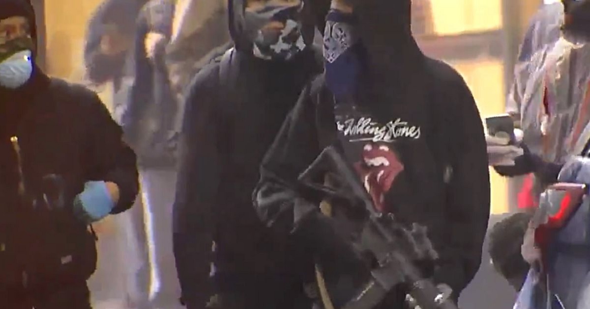 A masked rioter in Seattle holds a looted poliice rifle, pointed in a downward position.
