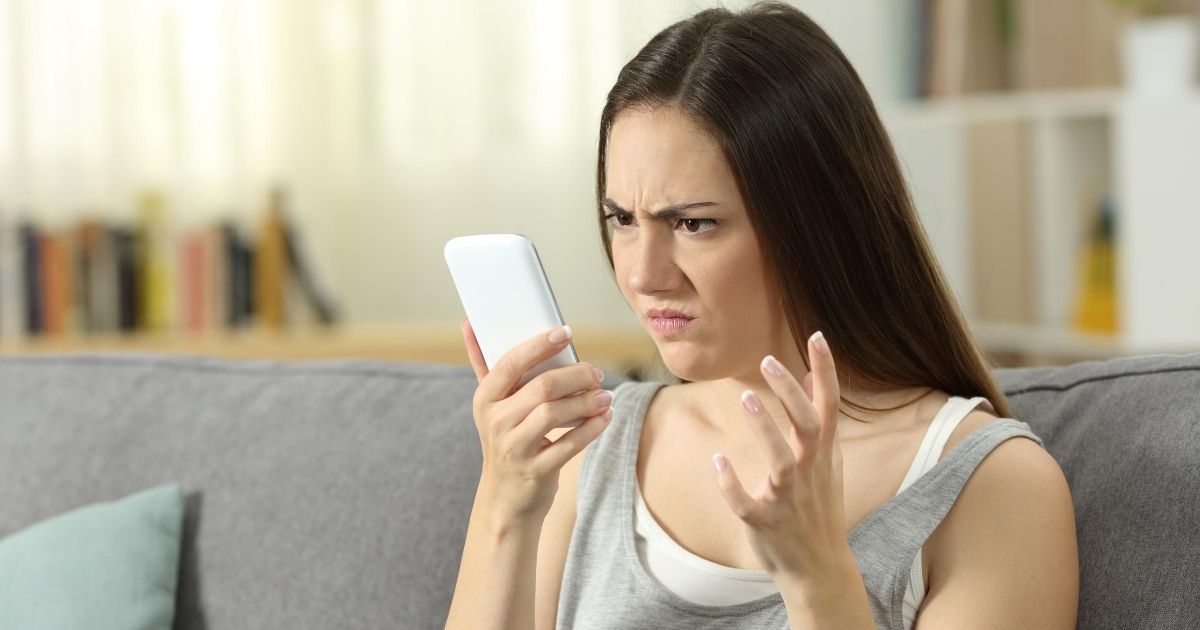 A woman on her couch looks at her cellphone with an angry look.