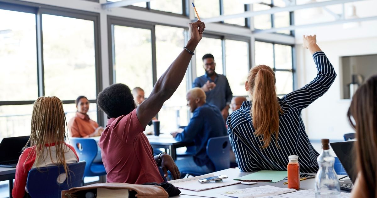 Students raise their hands at their desks in the above stock photo.
