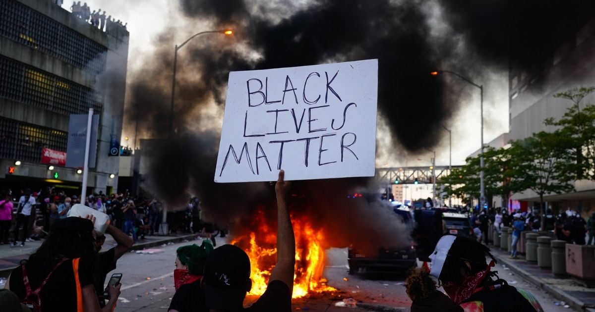 ATLANTA, GA - MAY 29: A man holds a Black Lives Matter sign as a police car burns during a protest on May 29, 2020 in Atlanta, Georgia. Demonstrations are being held across the US after George Floyd died in police custody on May 25th in Minneapolis, Minnesota.