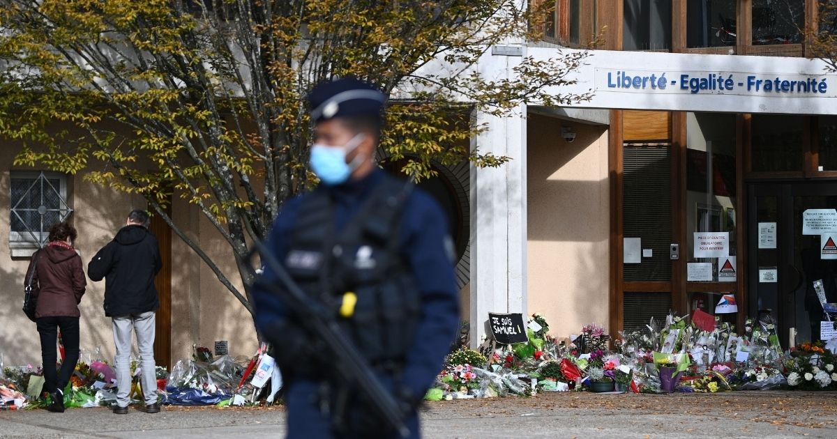 An officer stands guard Tuesday as people look at flowers placed outside the Bois d'Aulne secondary school in Conflans-Sainte-Honorine, France, in honor of slain history teacher Samuel Paty.