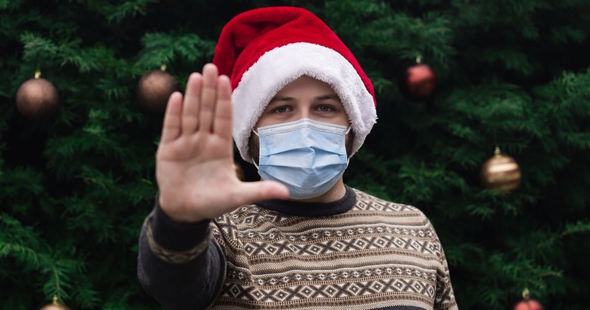 A man wearing a mask stands in front of a Christmas tree in the above stock image.