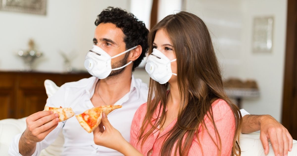 A mask-wearing couple eat pizza on the couch in the stock image above.