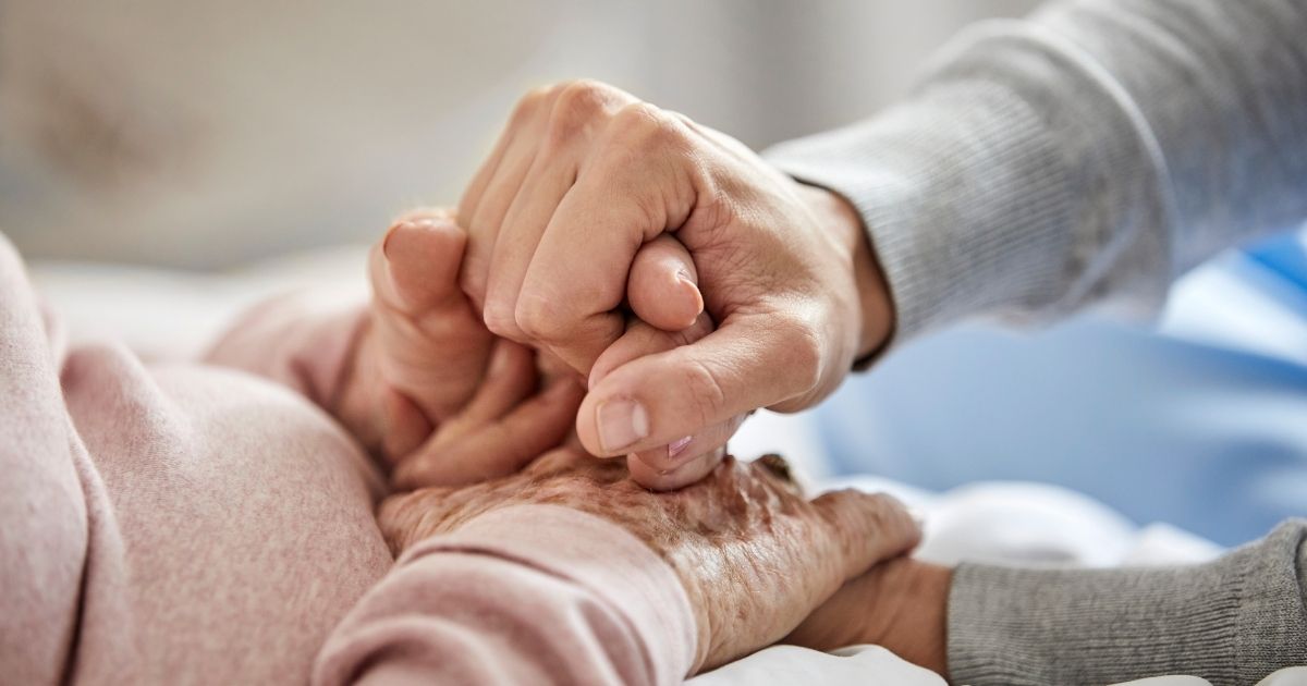 An elderly woman holds a caregiver's hand in the stock image above.