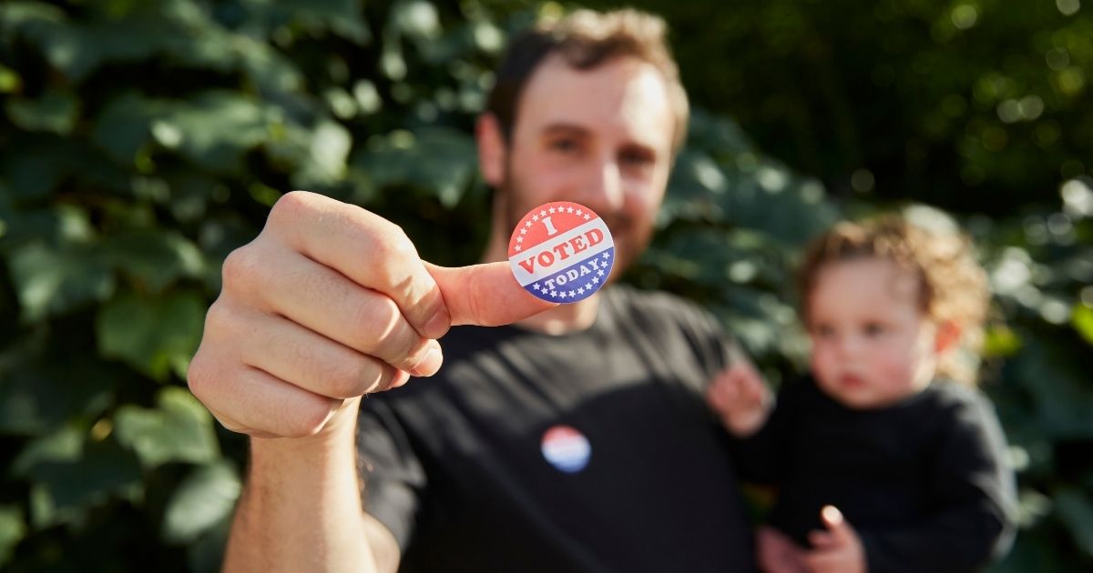 A father standing outside holding his baby, with an "I Voted Sticker" on his thumb in the above stock photo.
