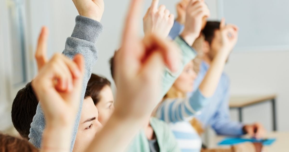 Students in a classroom raise their hands in the stock image above.