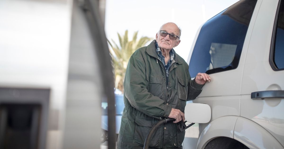 An older man grimaces while filling his vehicle at a gas pump.