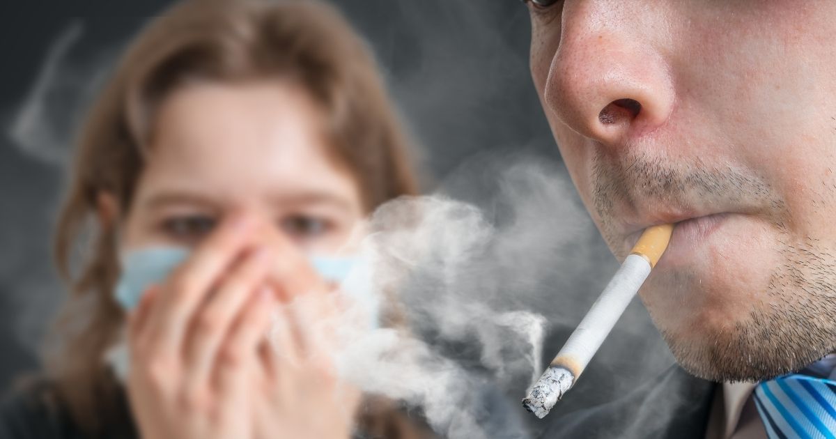 A man smokes a cigarette as a woman in a mask covers her face in the stock image above.