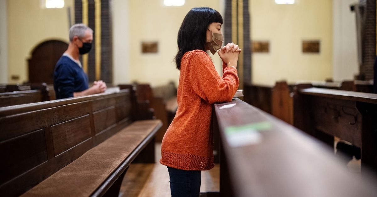 The above stock image shows a person praying with a face mask in a church.