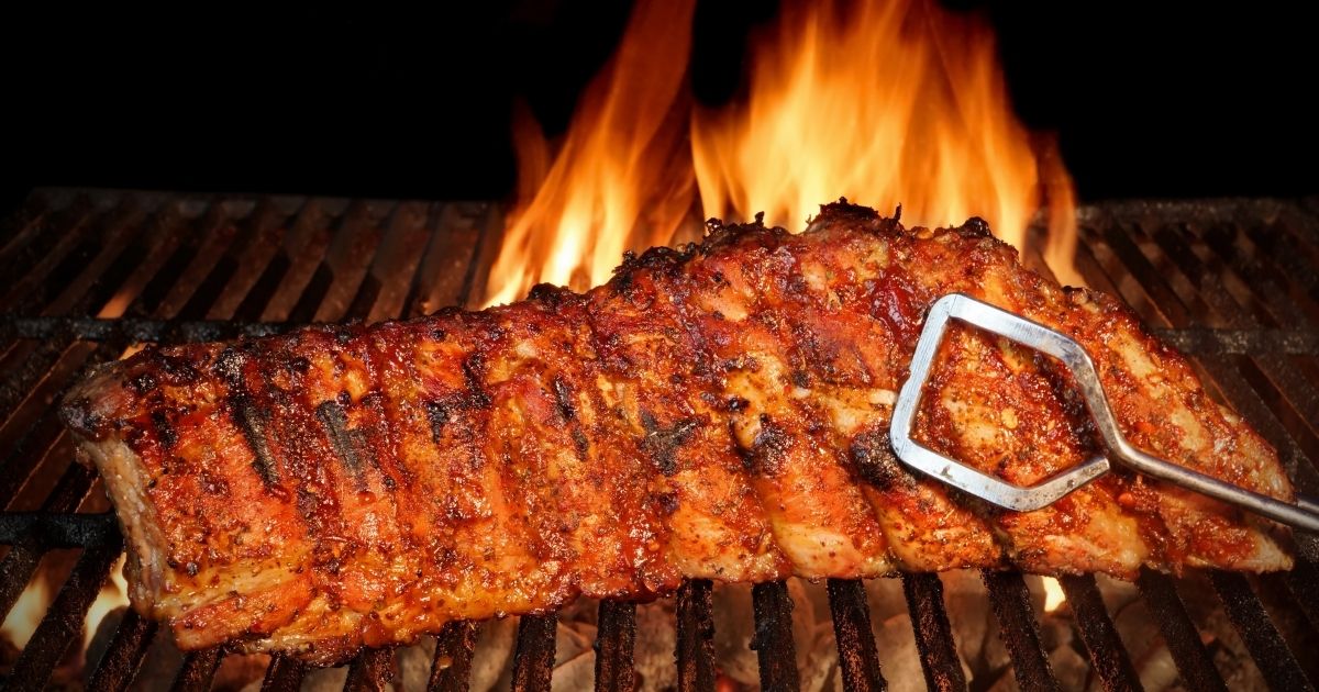 Baby back ribs cook on a flaming grill.