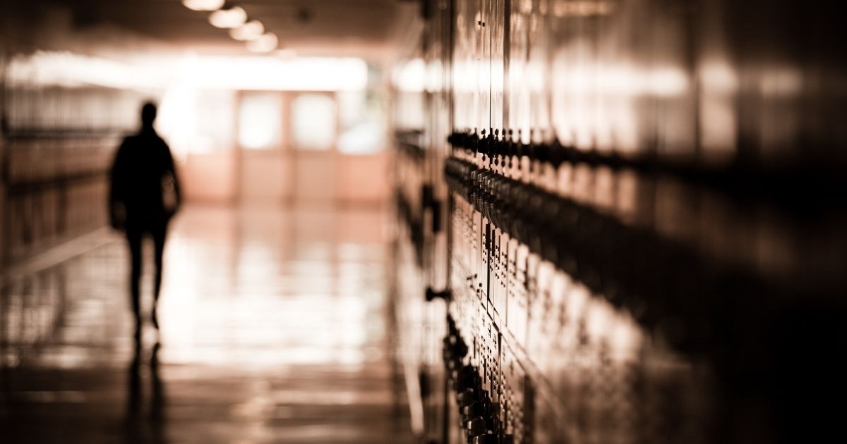 A high school student walks down a dark hallway in a public high school, silhouetted by daylight spilling in and reflecting off of the floor and lockers in the above stock photo.