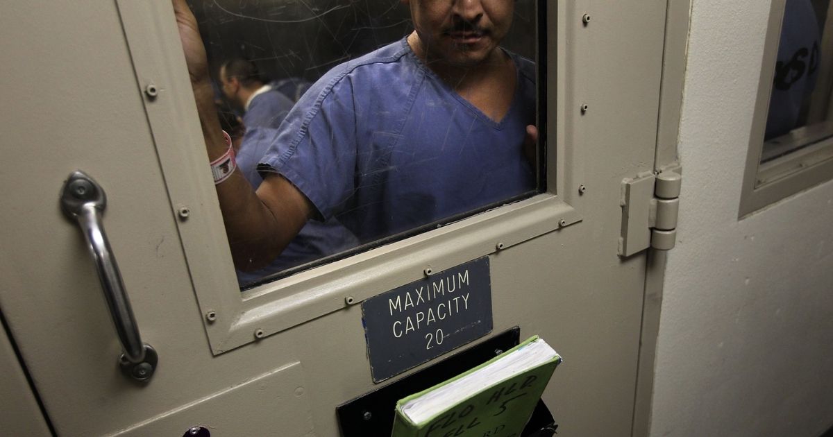 A detained migrant stands behind facility doors.