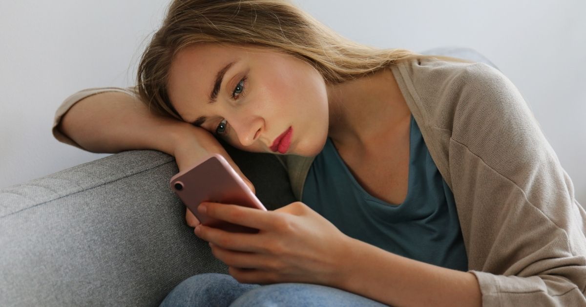 A young woman sits on a couch with a blank expression looking at her cellphone.