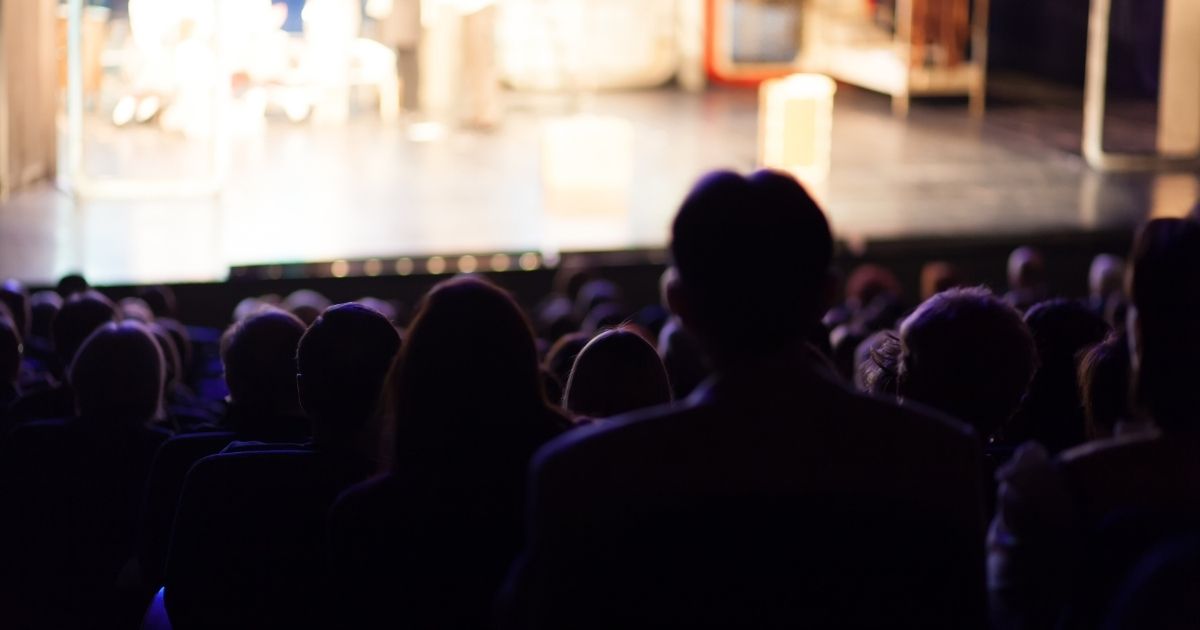 An audience in a theater watches a play in the above stock image. (