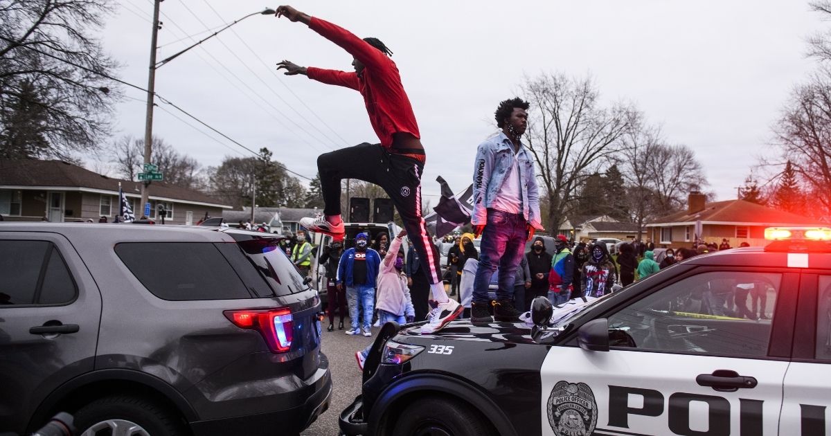 On man stands on the hood of a police car while another jumps to a nearby police vehicle.