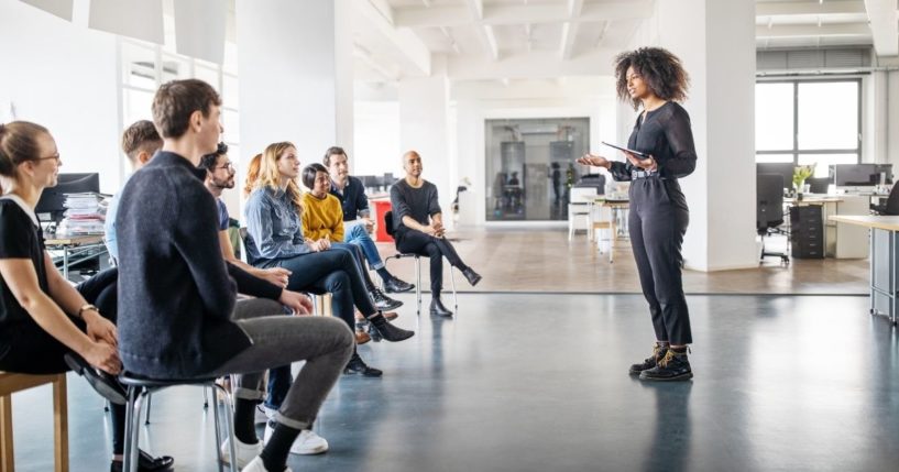 The above stock photo shows a woman speaking to a group of people.