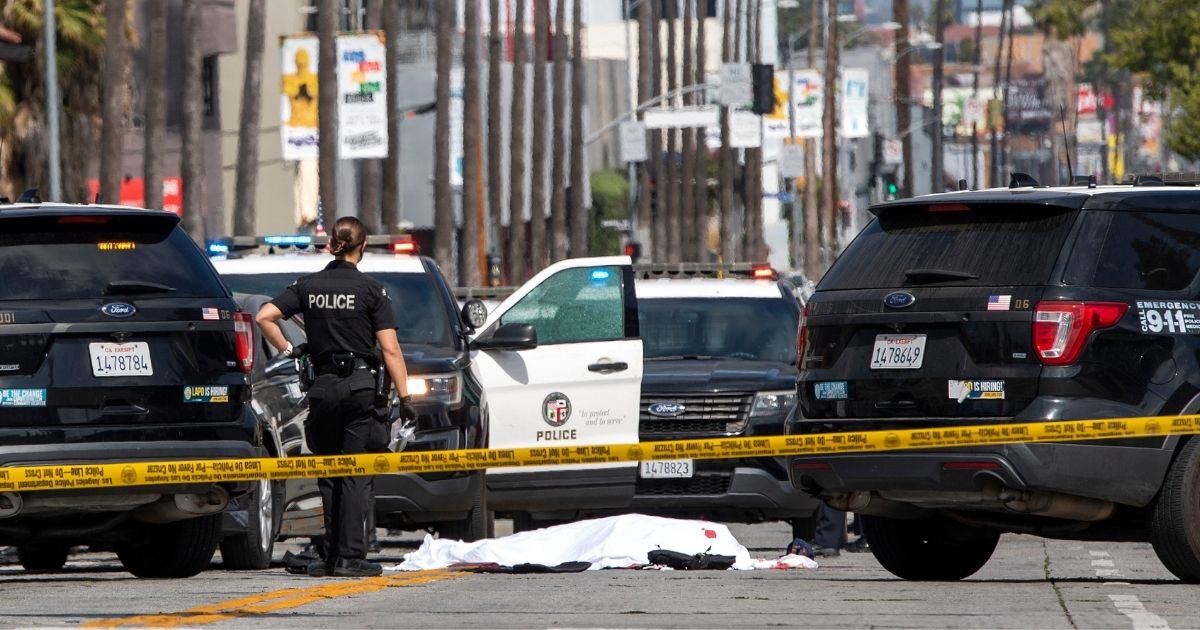 An LAPD police officer stands at the corner of Fairfax Avenue and Sunset Boulevard where a body covered in a white sheet lies on the pavement in Los Angeles on April 24, 2021.