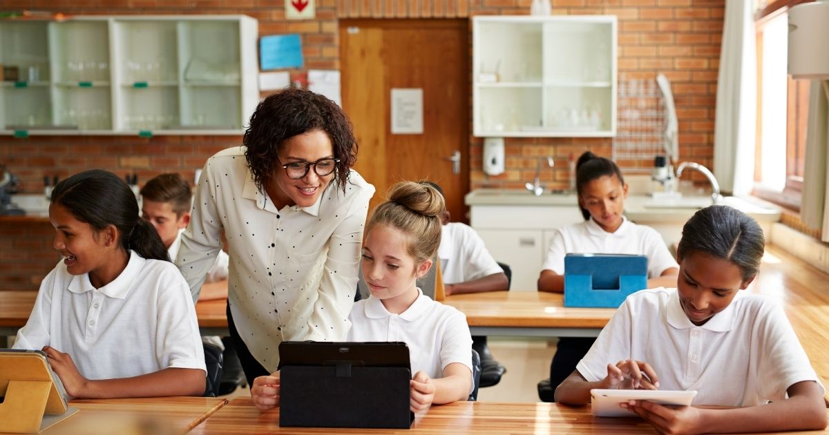 The above stock photo shows a teacher helping a student with a tablet in the classroom.