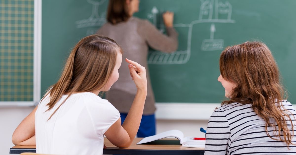 Two girls talk in class while a teacher writes on a blackboard.