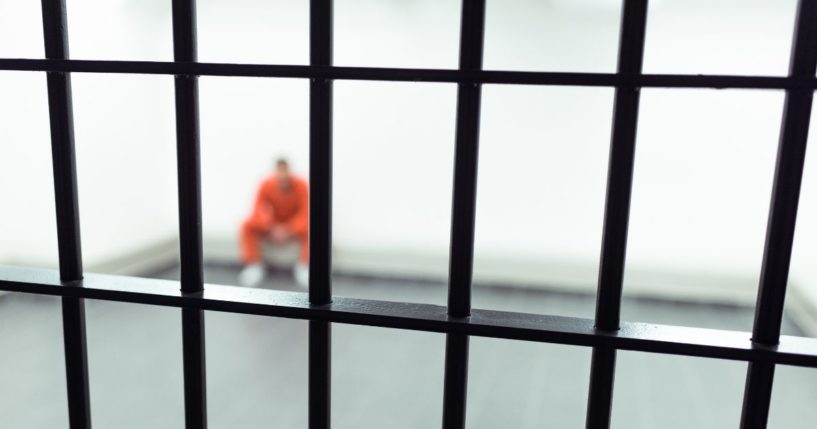 A male figure seated in a large jail cell.